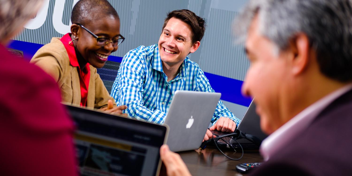 smiling adult student sitting at table with other adult students in front of their laptops